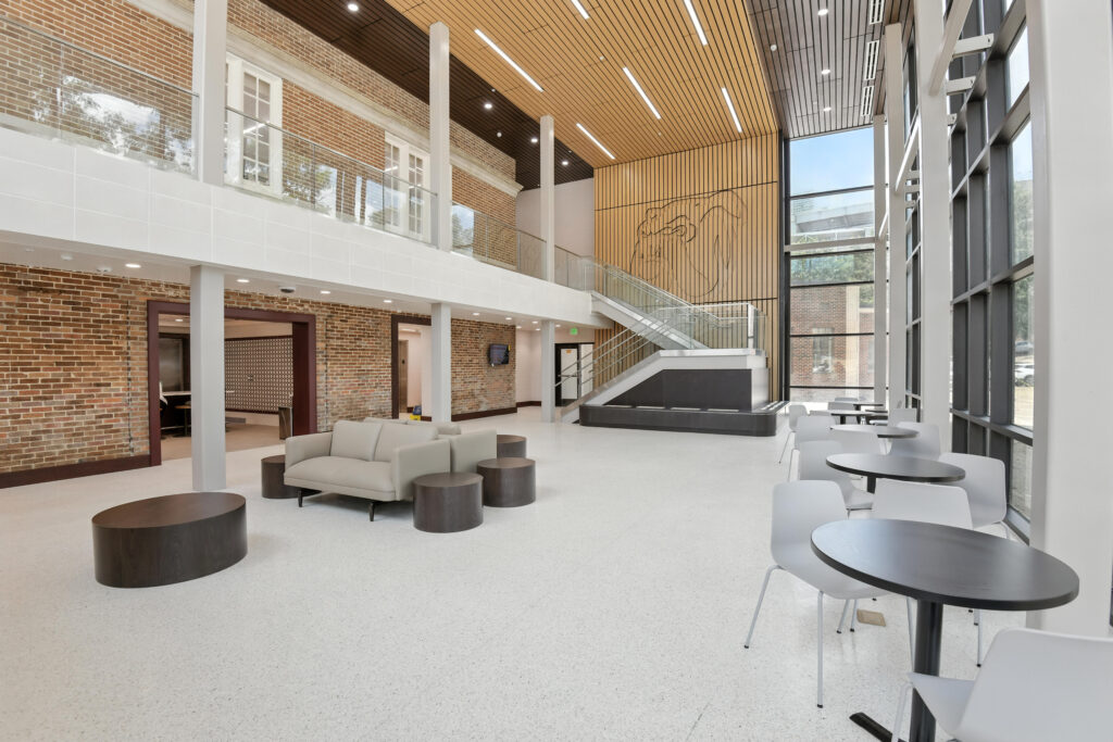 Modern two-story building lobby with a brick wall, seating area, staircase, and cafe tables.