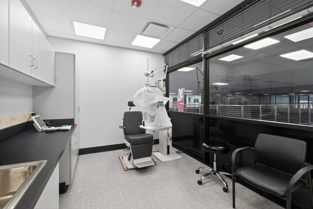 Medical examination room with a patient chair, a stool, and a side chair, viewed through a glass partition.