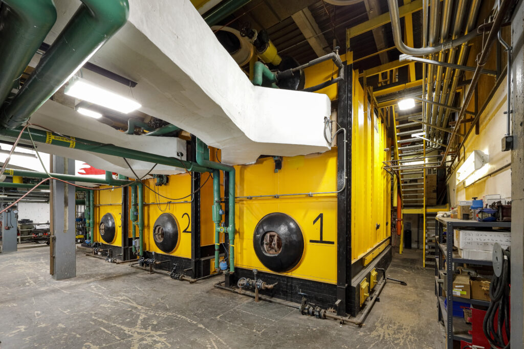Three large yellow industrial boilers numbered 1, 2, and 3 in a mechanical room, connected by overhead green pipes and white ductwork.