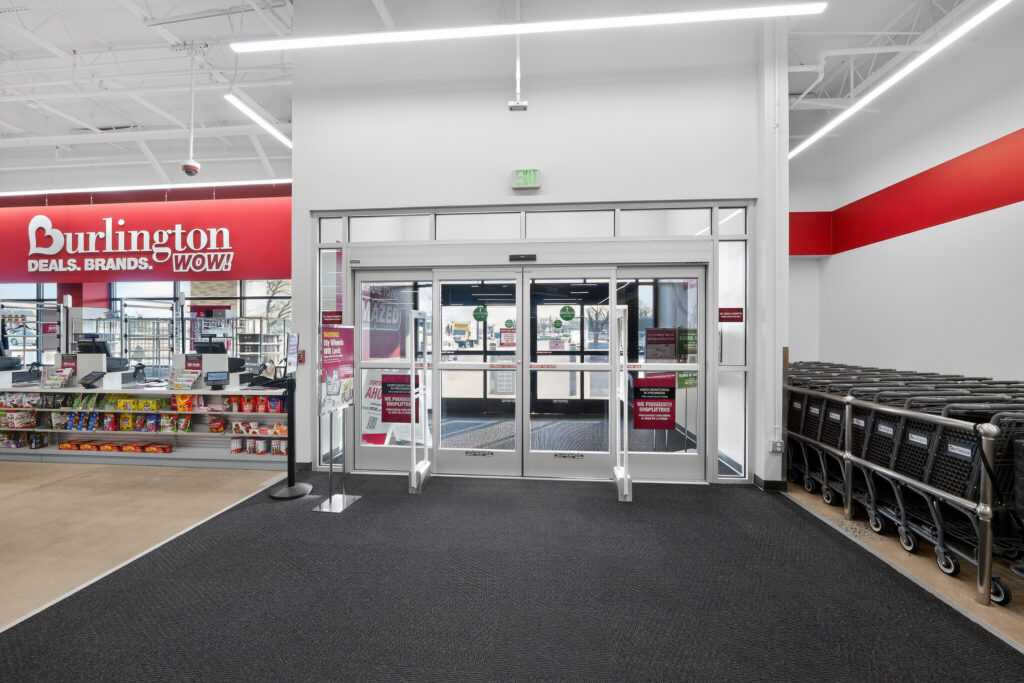 Entrance to a Burlington store with automatic glass doors, checkout counters to the left, and shopping carts lined up on the right.