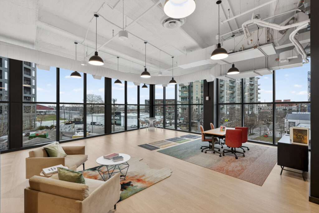 Open-concept office space featuring a lounge area with two armchairs and a small table, and a separate meeting area with a table and four chairs, surrounded by floor-to-ceiling windows.