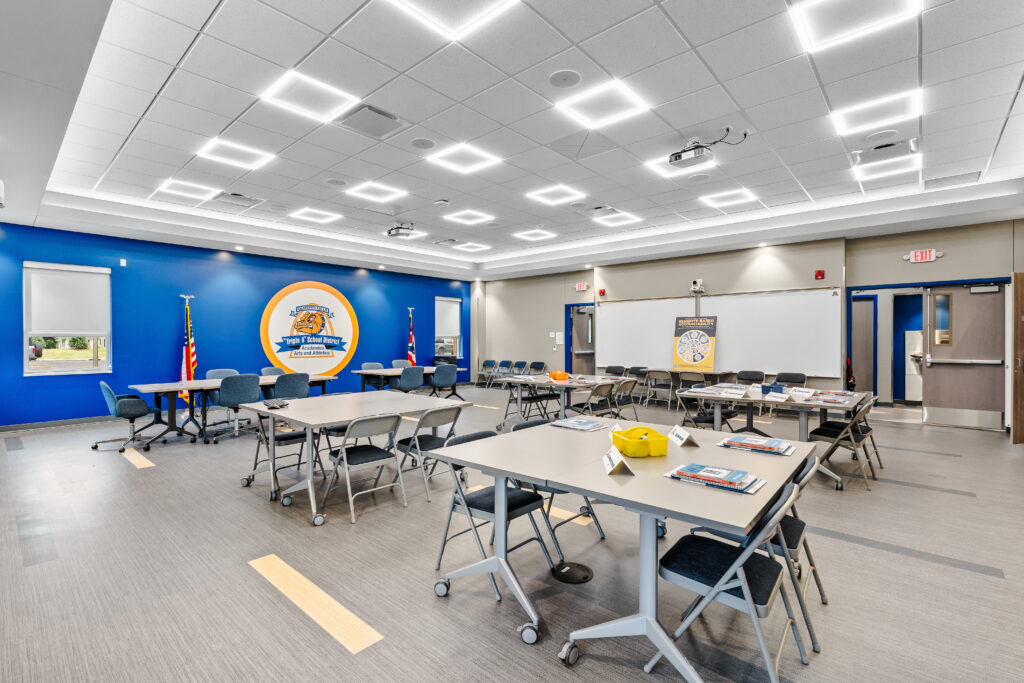 A multi-purpose room set up with rows of tables and chairs, featuring a blue accent wall with a district logo and a dry-erase board at the front.