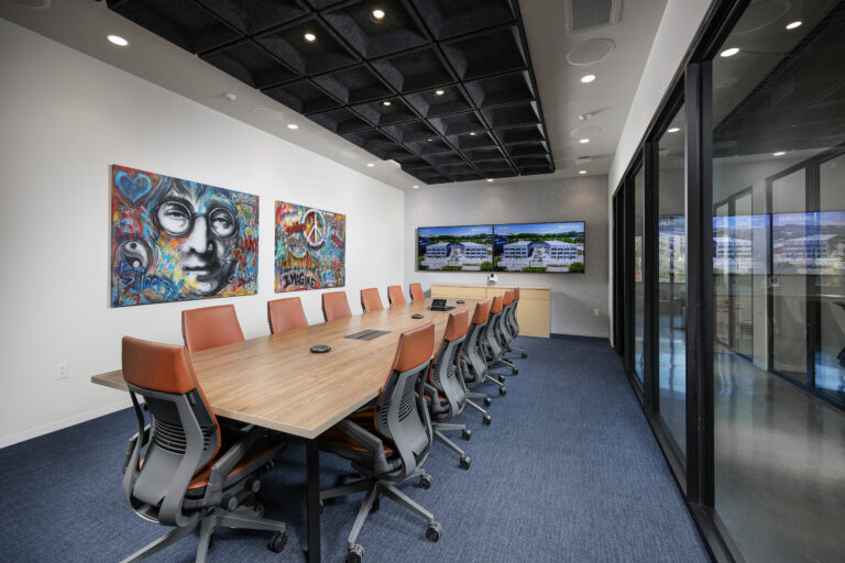 Conference room with a long wood-top table, orange office chairs, two wall-mounted televisions, and two colorful paintings on the wall.