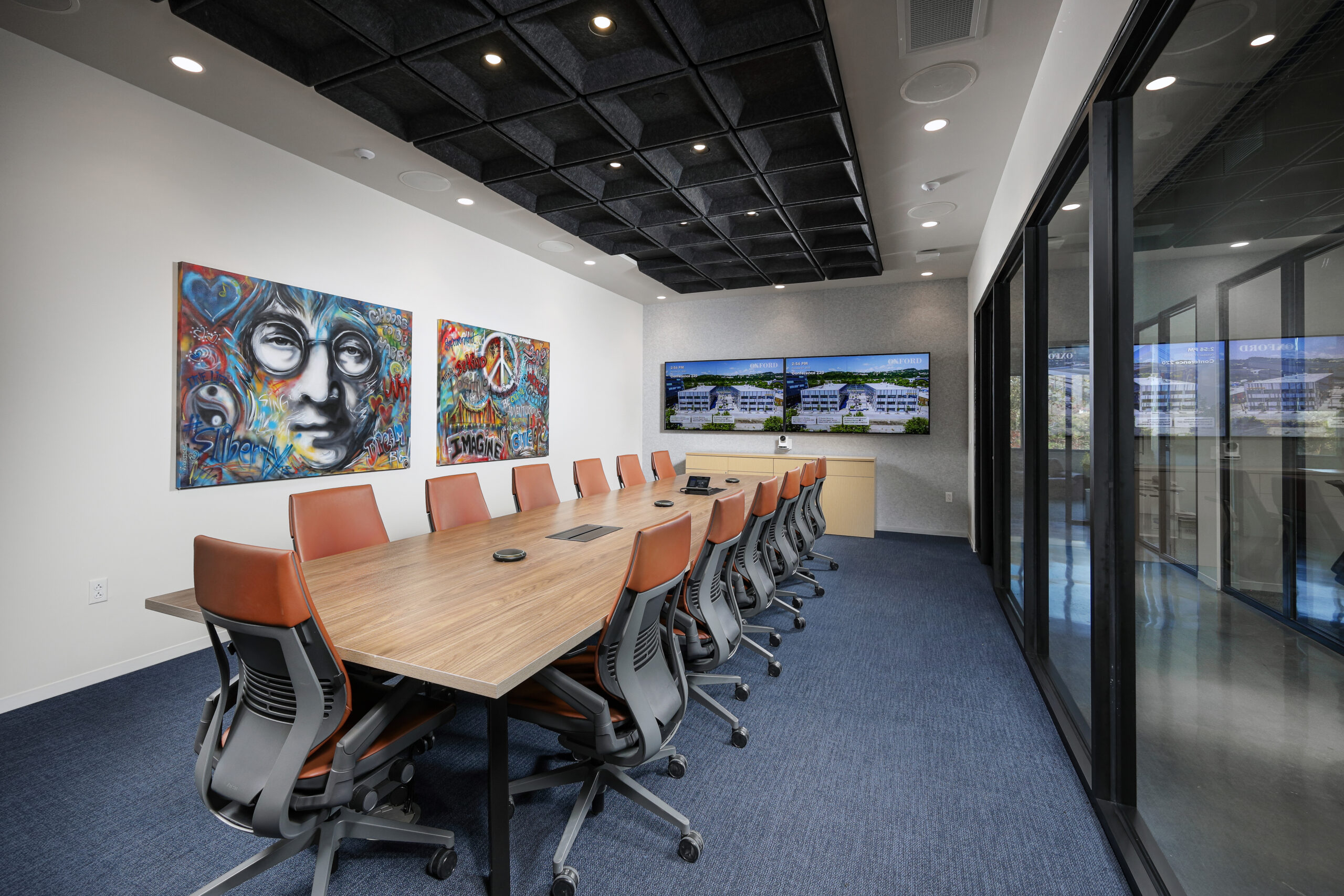 Conference room with a long wood-top table, orange office chairs, two wall-mounted televisions, and two colorful paintings on the wall.