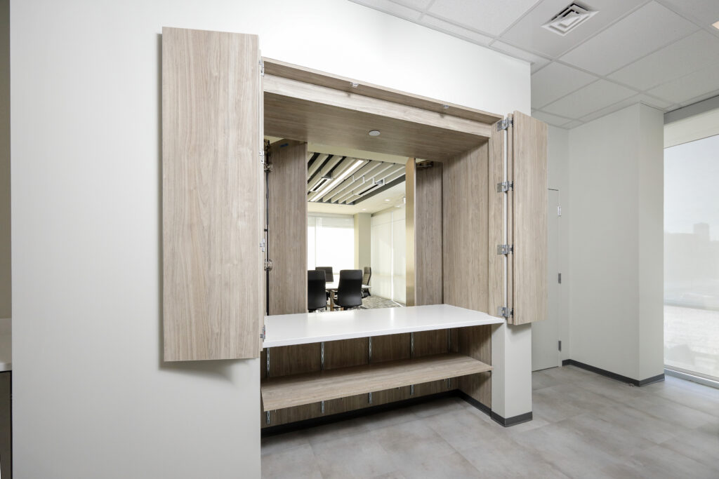 A wall-mounted desk unit with light wood cabinetry and an open work surface, showing a view of a conference room behind it.