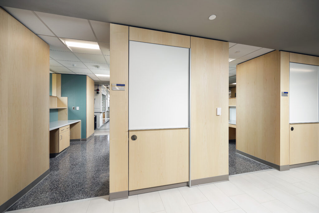 A modern office interior featuring light wood partitions, a white dry-erase board, and a corridor leading to a workspace with teal walls.