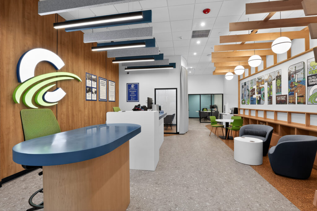 Modern office lobby with a blue reception desk, a large green and white logo on the wall, a seating area with lounge chairs, and a graphic wall mural.
