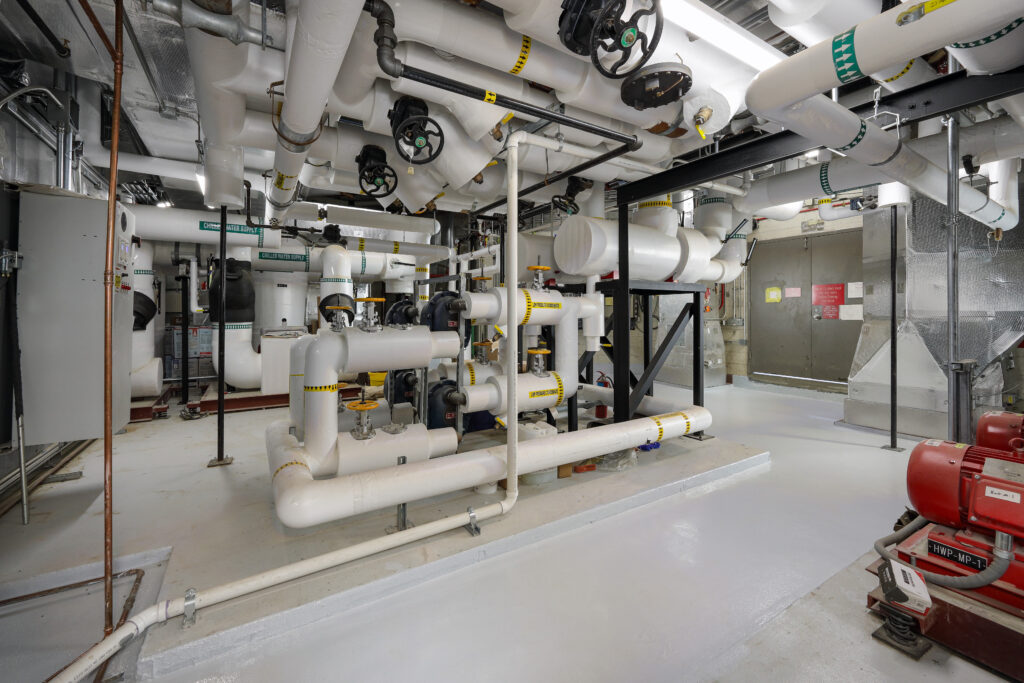 Industrial mechanical room featuring a network of insulated white pipes with valves and directional arrows, and a red pump on the floor.