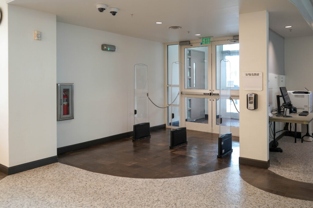 Building exit with security pedestals, a fire extinguisher cabinet, and a nearby desk with a computer.