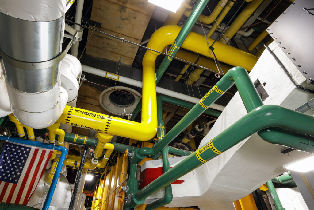 A network of pipes in a mechanical room, featuring yellow “high pressure steam” pipes and green “boiler feed water” pipes, with an American flag visible in the lower left.