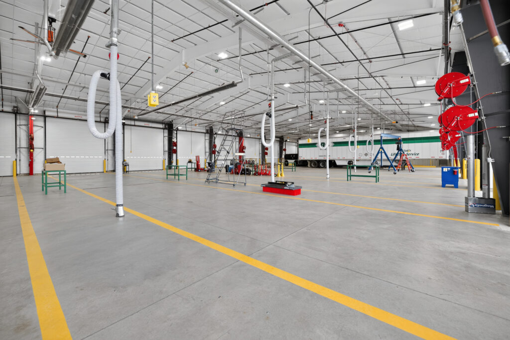 Spacious industrial garage with overhead ventilation hoses, yellow floor markings, a white trailer, and various maintenance equipment.