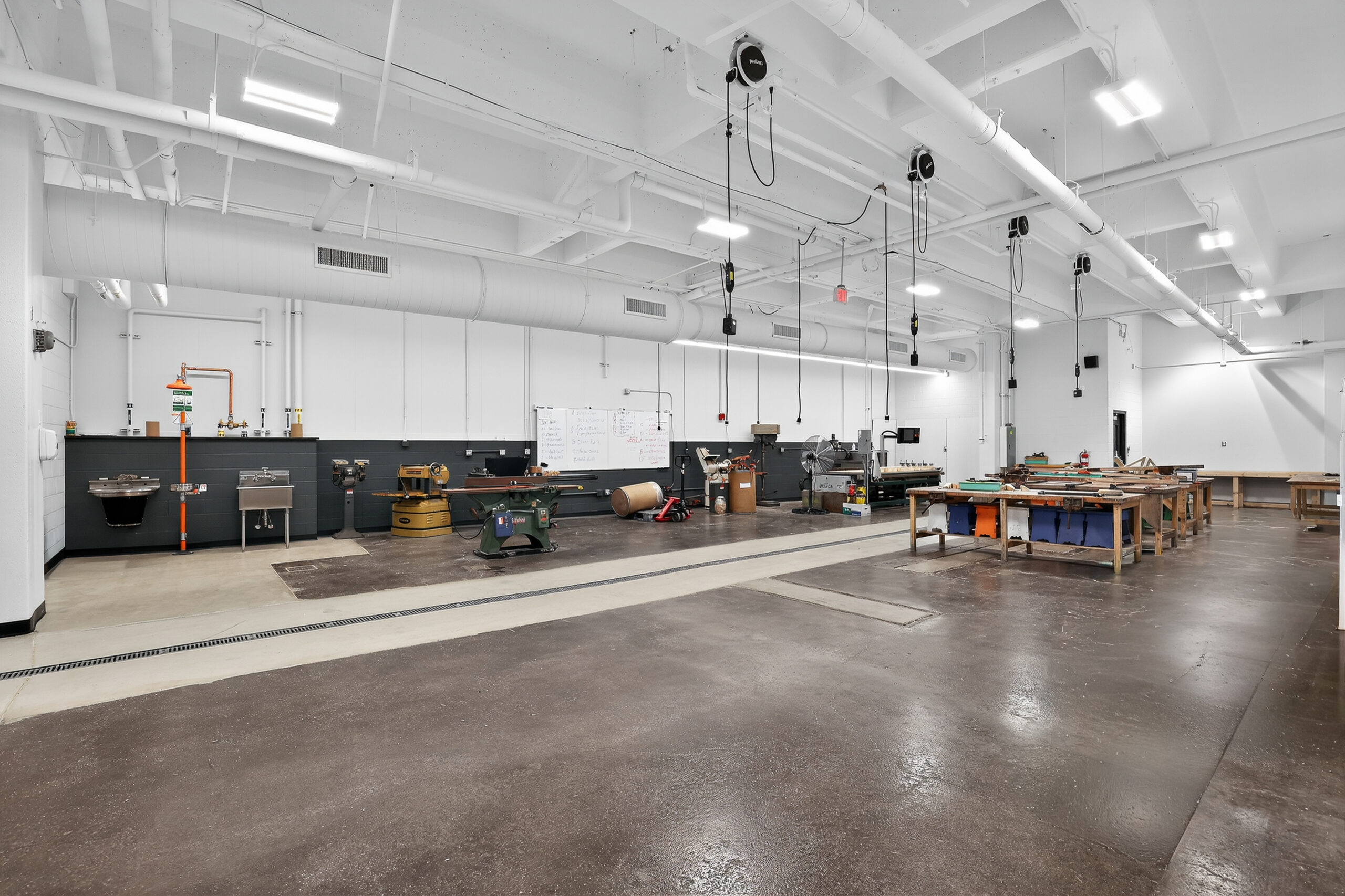 A workshop with a concrete floor, equipped with woodworking machinery, a sink, and several workbenches.