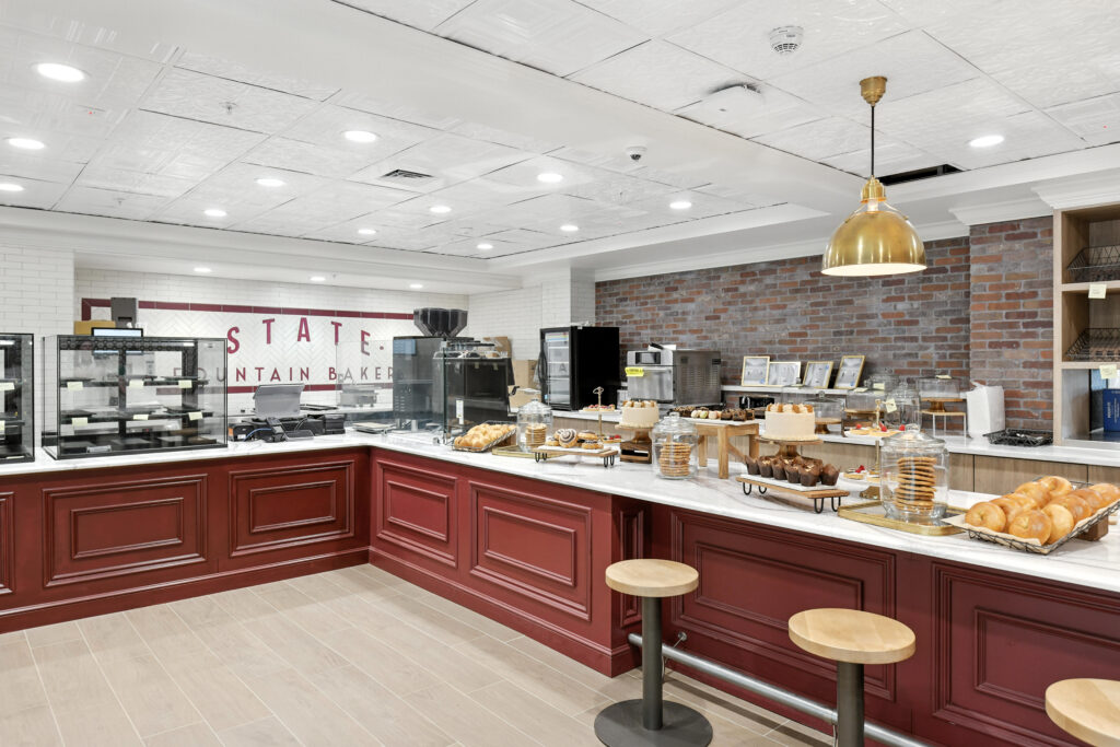 A bakery counter with a display of pastries, cakes, and cookies, featuring a red base and white marble top with the words “State Fountain Bakery” on the wall behind.