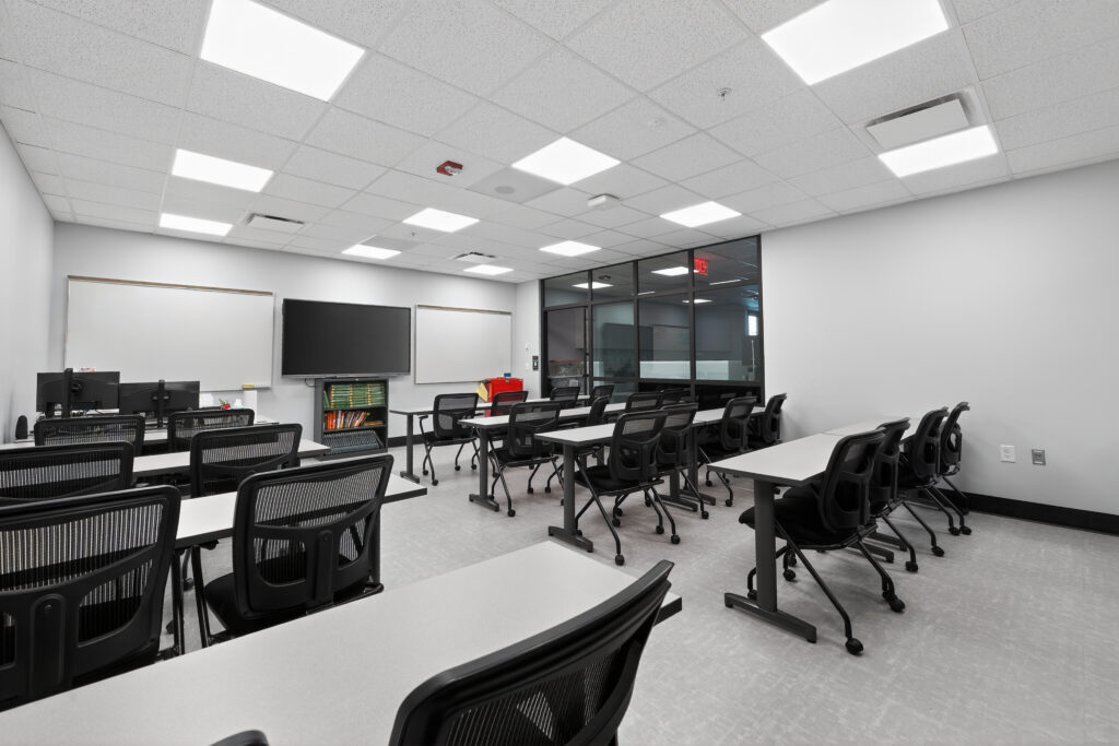 A modern classroom with rows of desks, black mesh chairs, a large wall-mounted monitor, and whiteboards.
