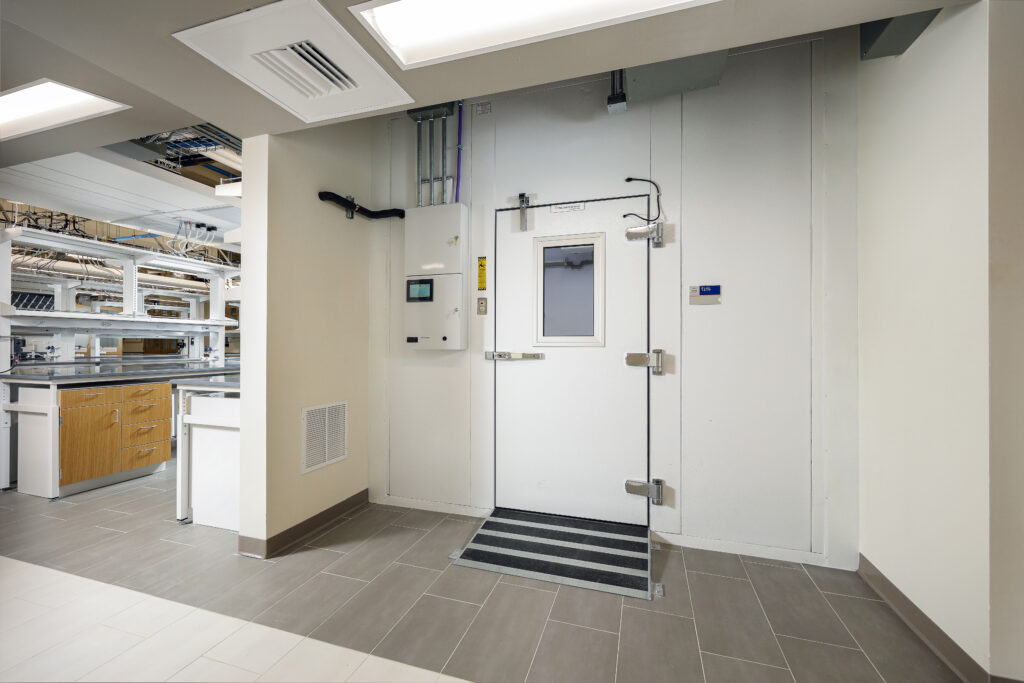 A white industrial walk-in door with a small window, latch, and ramp, situated in a laboratory setting.