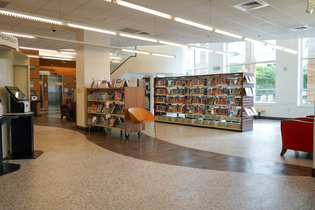 The interior of a library with a self-checkout station, a mobile book display, and rows of bookshelves.