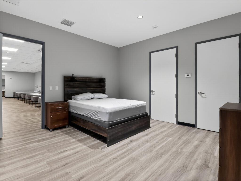 A bedroom with a queen-sized bed, a wooden nightstand, and two white doors, looking into a hallway lined with additional beds.