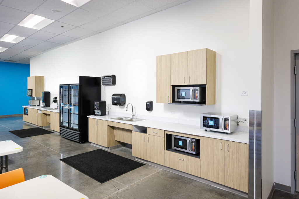 A modern office breakroom kitchen featuring light wood cabinets, a sink, a refrigerator, and three built-in microwaves.
