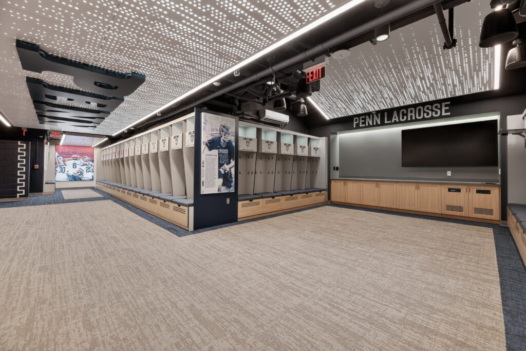 A Penn Lacrosse locker room featuring rows of lockers, a wall-mounted display of a player, a flat-screen television, and a lighted ceiling with a dot pattern.