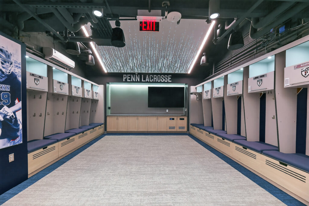Modern Penn Lacrosse locker room with two rows of lockers, a central bench area, and a wall-mounted television.