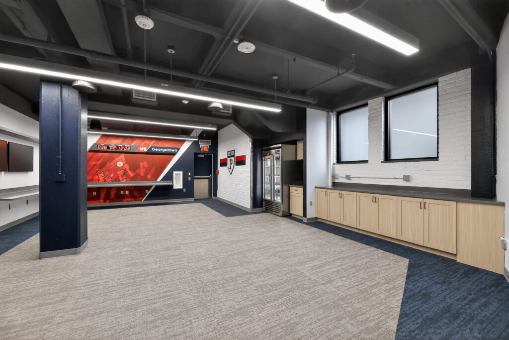An interior view of a room with a large screen displaying “Georgetown” vs. “Penn,” a small refrigerator, light wood cabinets, and gray carpet.