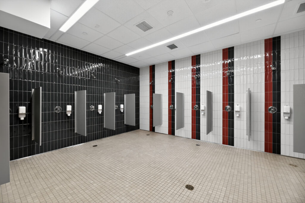 A locker room shower area featuring multiple stalls with gray partitions against tiled walls of black, white, and red.