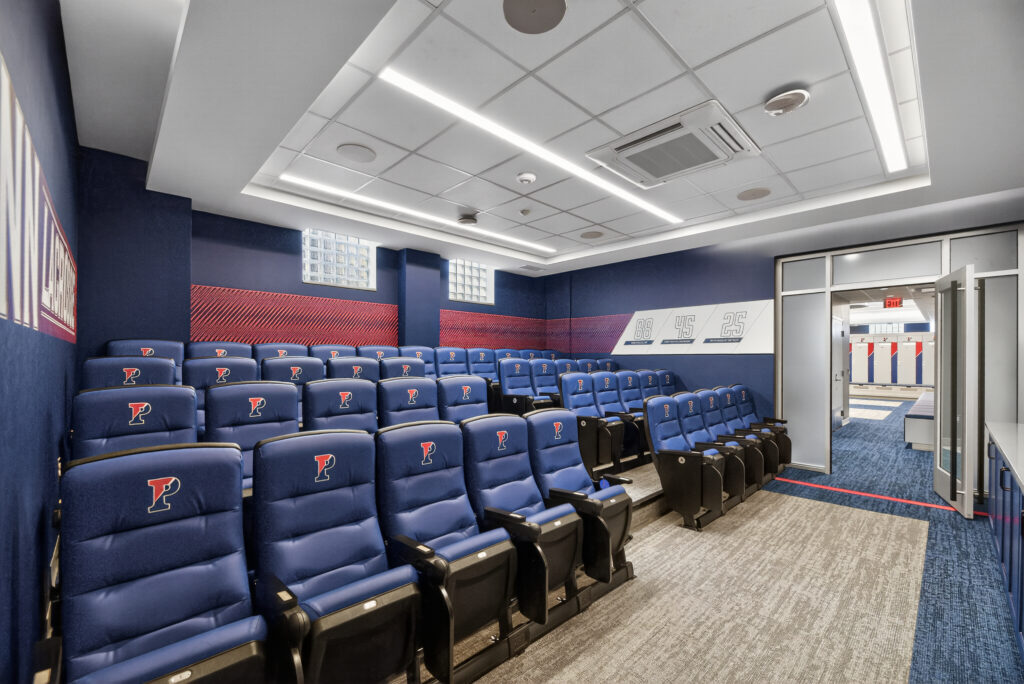 A tiered room with rows of blue leather theater seats, each featuring a letter P logo, leading into an adjacent locker room.