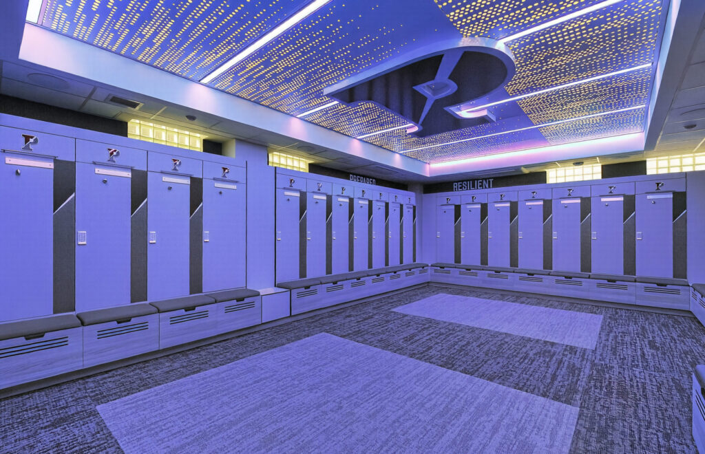 Locker room with rows of gray lockers and benches, featuring a backlit ceiling pattern and the word “RESILIENT” on the back wall.