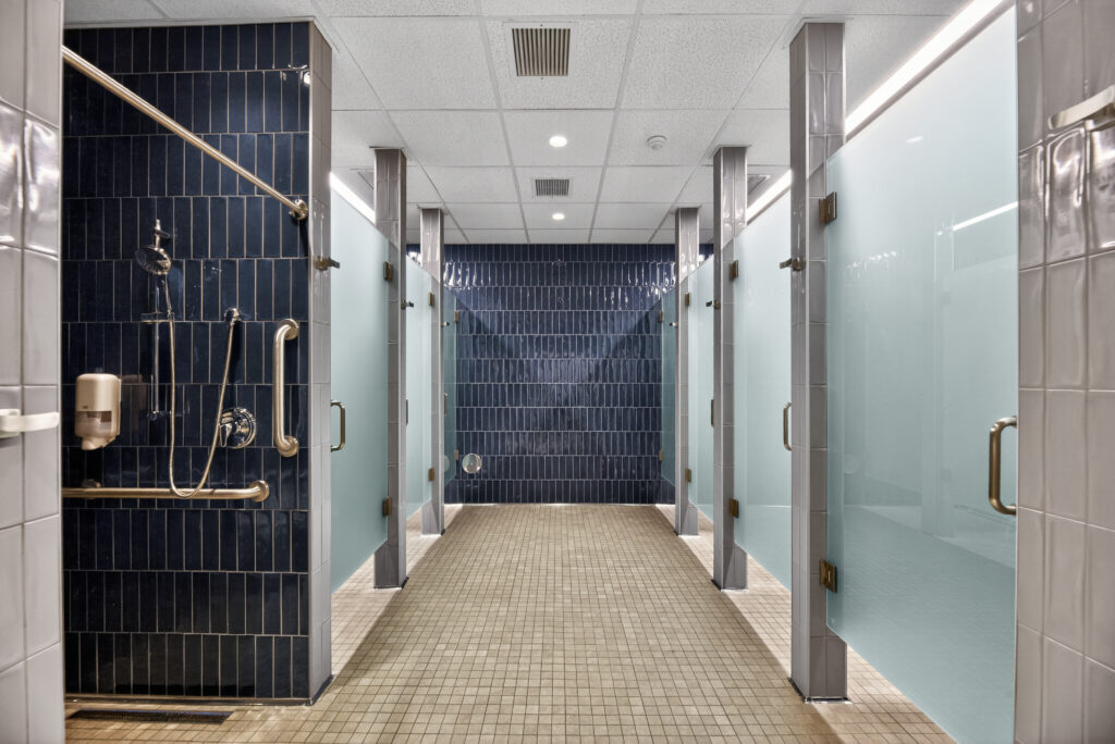 A row of shower stalls with blue tiled walls and frosted glass doors, featuring grab bars and mounted soap dispensers.