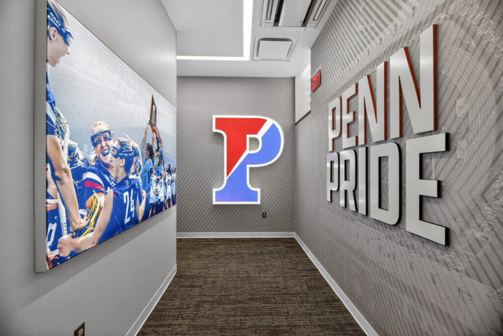 A hallway featuring a large photo of a lacrosse team, a backlit “P” logo on the wall, and three-dimensional “PENN PRIDE” lettering.