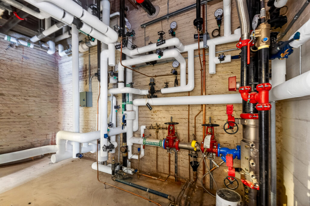 A utility room showing a network of white insulated pipes, pressure gauges, and red-handled valves against a brick wall.