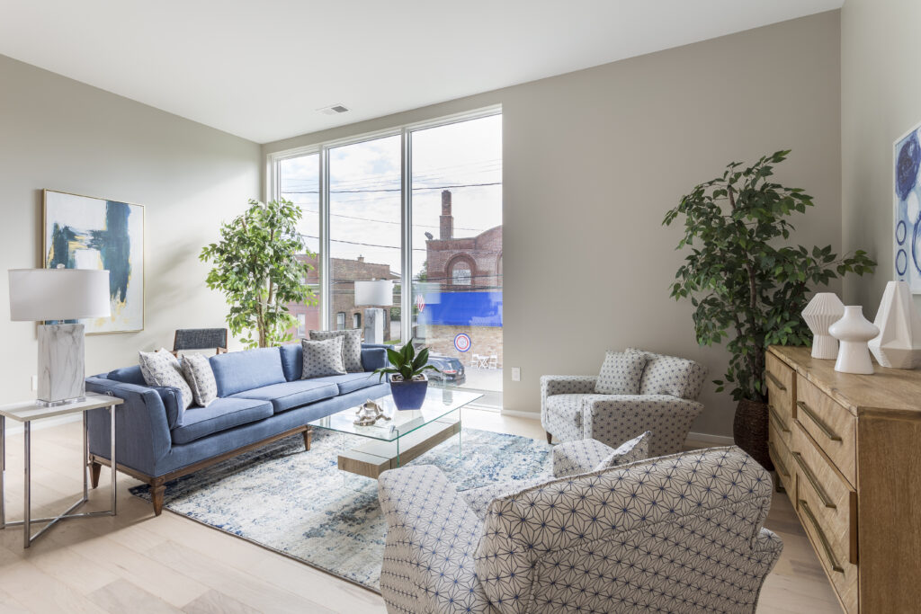 A blue sofa, two patterned armchairs, and a glass coffee table in a sunlit living room with floor-to-ceiling windows.