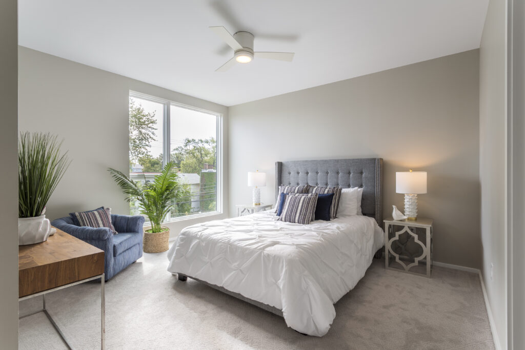 A bedroom featuring a gray tufted headboard, a white bed, two bedside tables with lamps, a blue armchair, and a wooden desk.