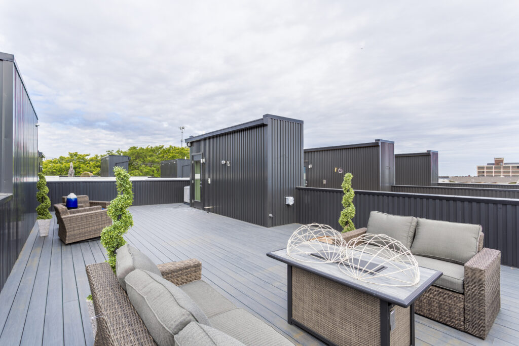 Rooftop terrace with gray decking, dark corrugated metal structures, and wicker lounge furniture with gray cushions.