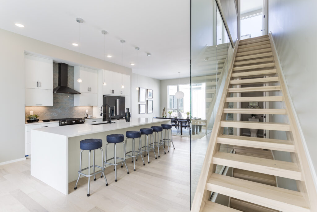 Modern kitchen with a white island and blue stools, viewed next to an open wooden staircase with a glass railing.