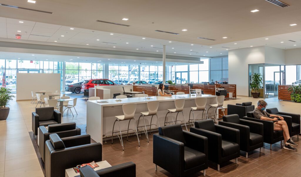 A spacious car dealership waiting area with black armchairs, a long high-top table with stools, and a customer working on a laptop.