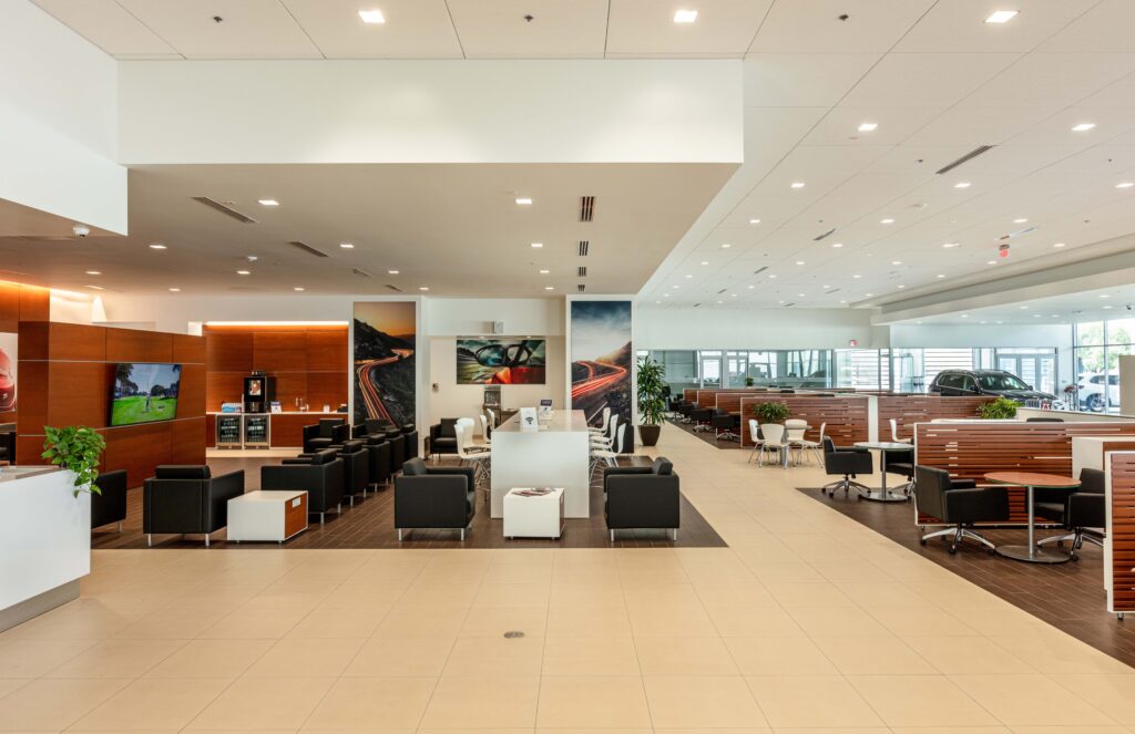 Modern car dealership waiting area with rows of black chairs, tables, and wooden partitions.