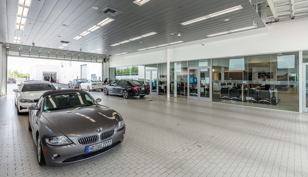 Several BMW cars parked inside a bright, modern dealership showroom with glass-walled offices visible in the background.