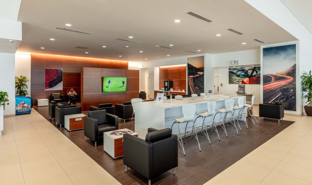 Modern lounge area with a long high-top table, white stools, black armchairs, and wall-mounted televisions.