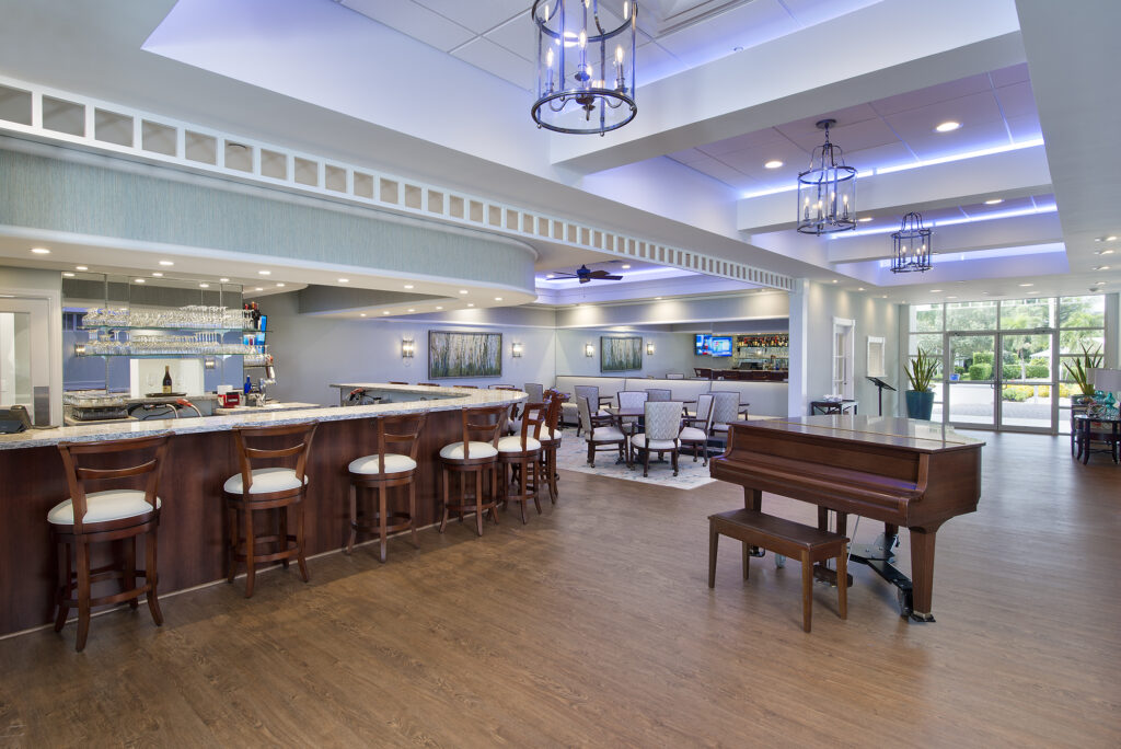 A hotel bar area with a wooden grand piano in the foreground, a curved bar with high stools, and tables with chairs in the background.