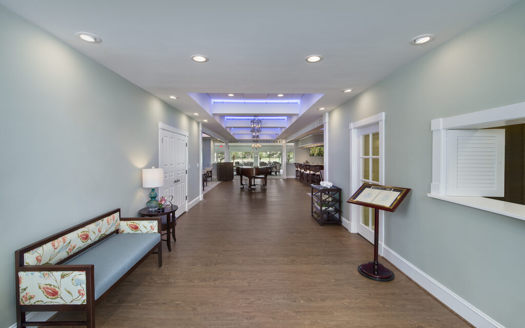A brightly lit hallway leading to a dining area featuring a grand piano and bar seating, with a floral bench in the foreground.