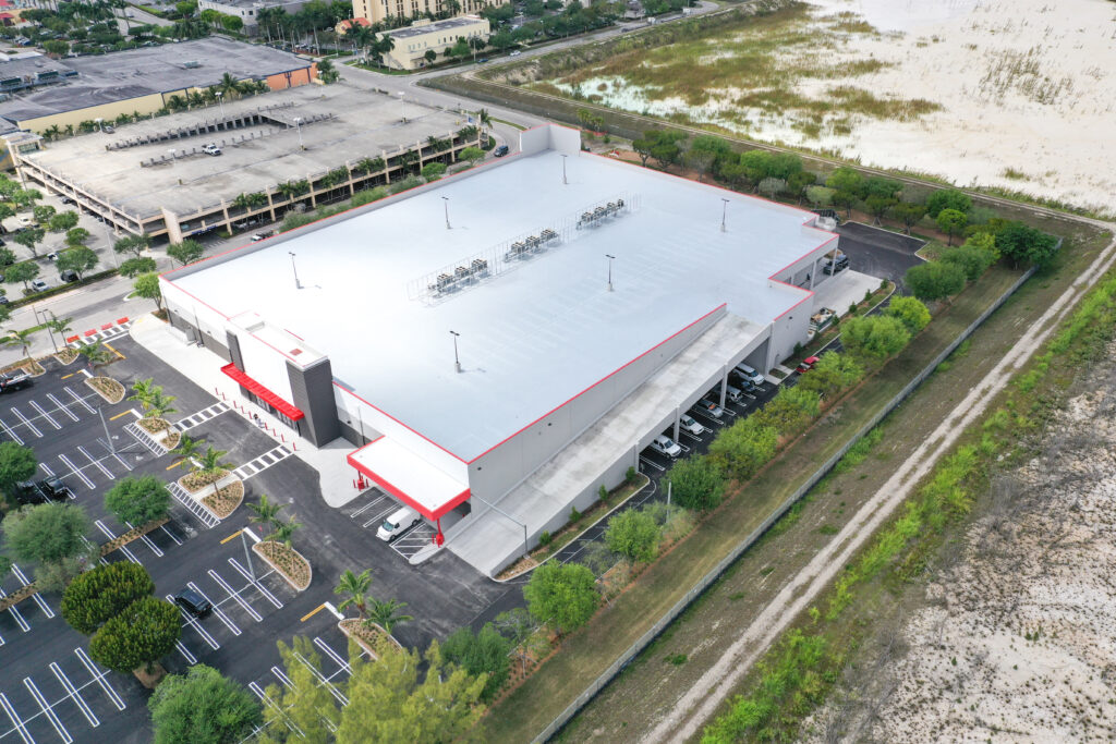 Aerial view of a large commercial building with a grey roof, red trim, and an adjacent parking lot.