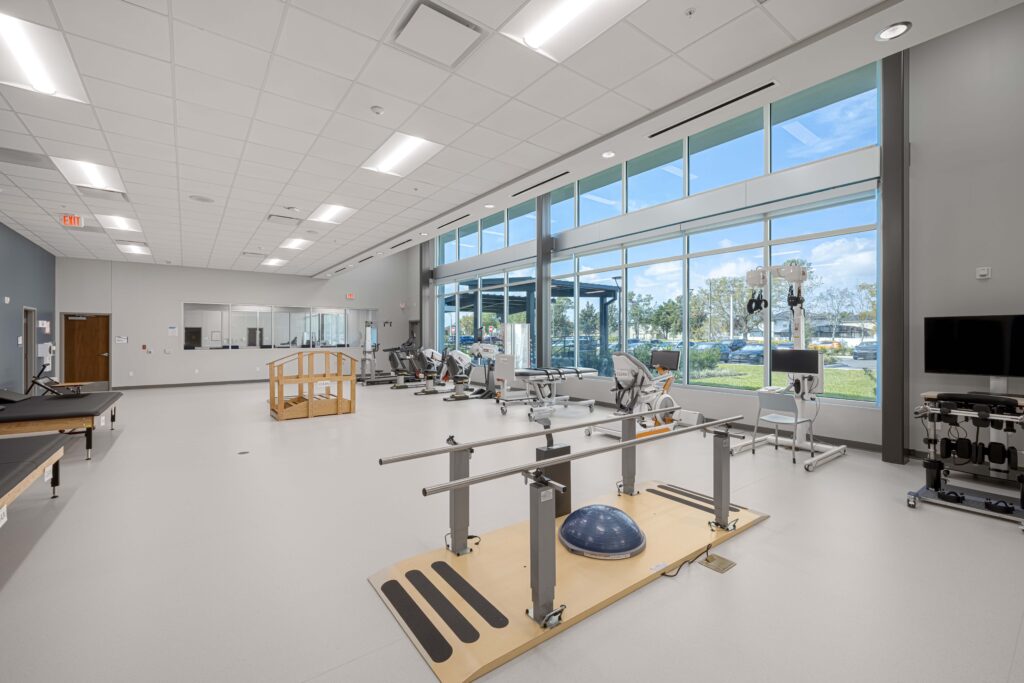A physical therapy clinic with parallel bars, a balance ball, exercise equipment, and wooden stairs in a large, window-lined room.