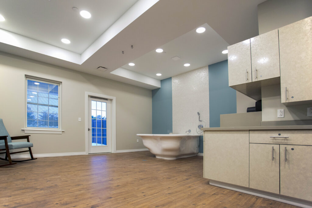 A room featuring a white freestanding bathtub against a teal and white tiled wall, a light-colored kitchen cabinet, a rocking chair, a window, and a glass-paned door.