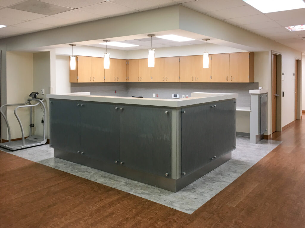 A reception desk with a gray paneled front, light wood cabinets, and four pendant lights, with an elliptical trainer in the corner.