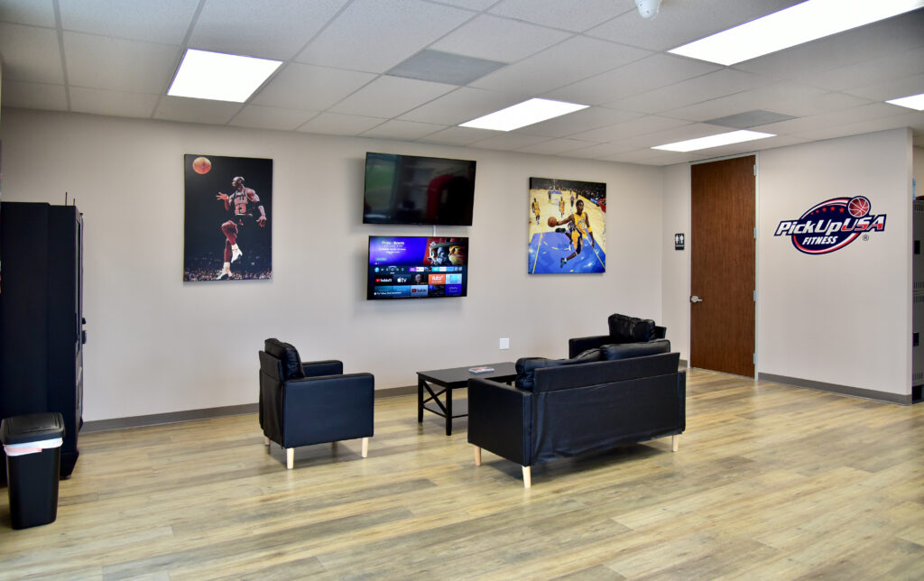 Waiting area with two black armchairs, a black sofa, a coffee table, and two televisions mounted on a wall decorated with basketball posters and a PickUp USA Fitness logo.