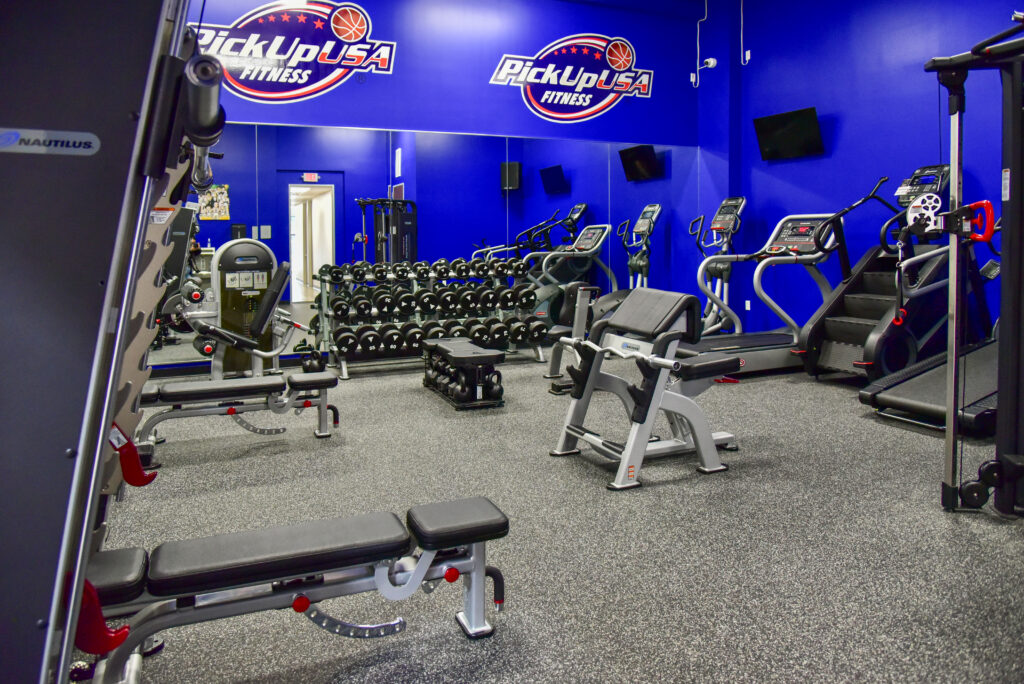 Interior of a PickUp USA Fitness gym with weight benches, a dumbbell rack, and cardio machines against a blue wall with the gym logo.