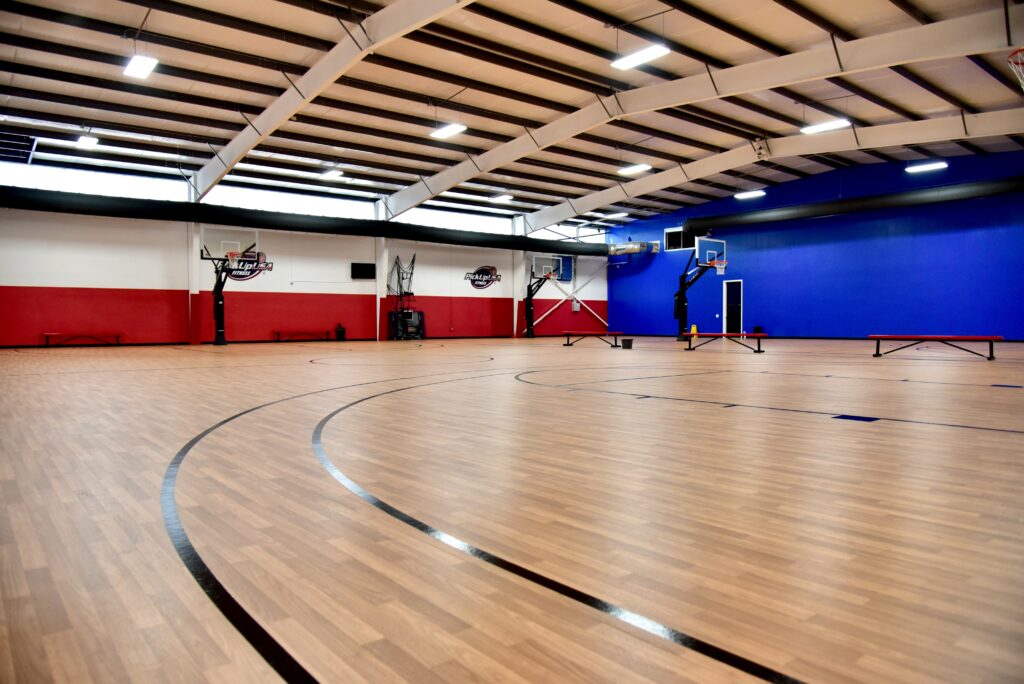 Indoor basketball court with wooden flooring, multiple hoops, and red and blue wall sections.