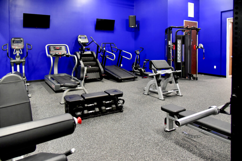 A gym with a blue wall featuring cardio machines, weight benches, and a multi-station weight rack on a grey floor.