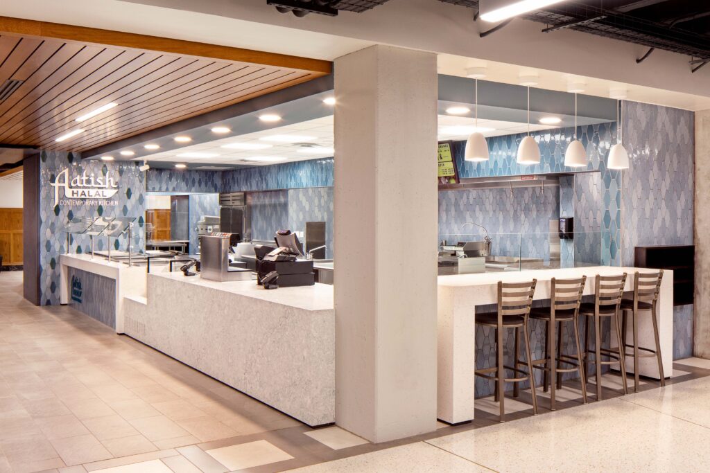 A modern restaurant interior with a long service counter, blue hexagon-tiled walls, and a high-top seating area with four stools.